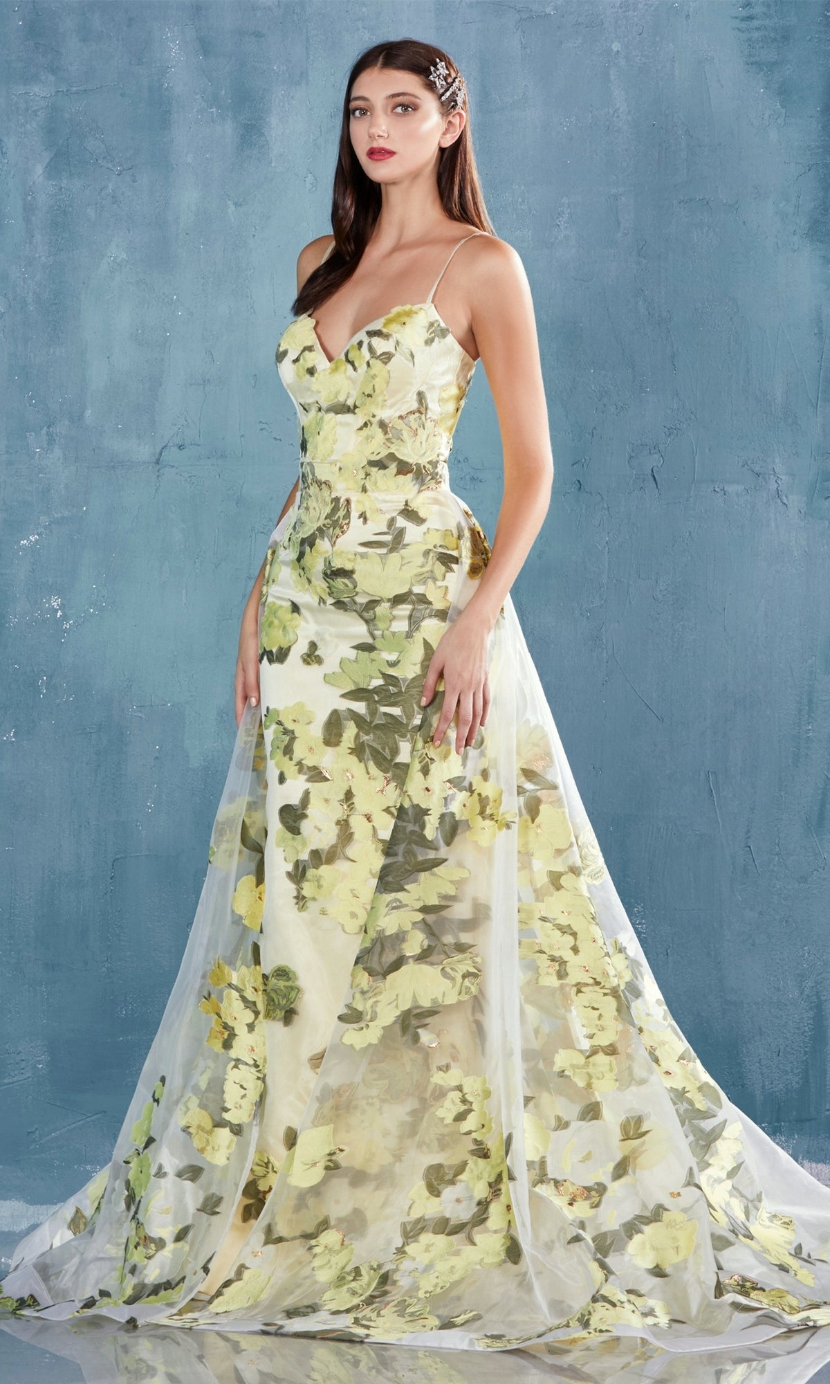 Against a textured blue background, a woman wears the Lemon Yellow Floral-Print Long Prom Dress A0770—a floor-length, sleeveless gown featuring a green floral pattern and sheer overlay.